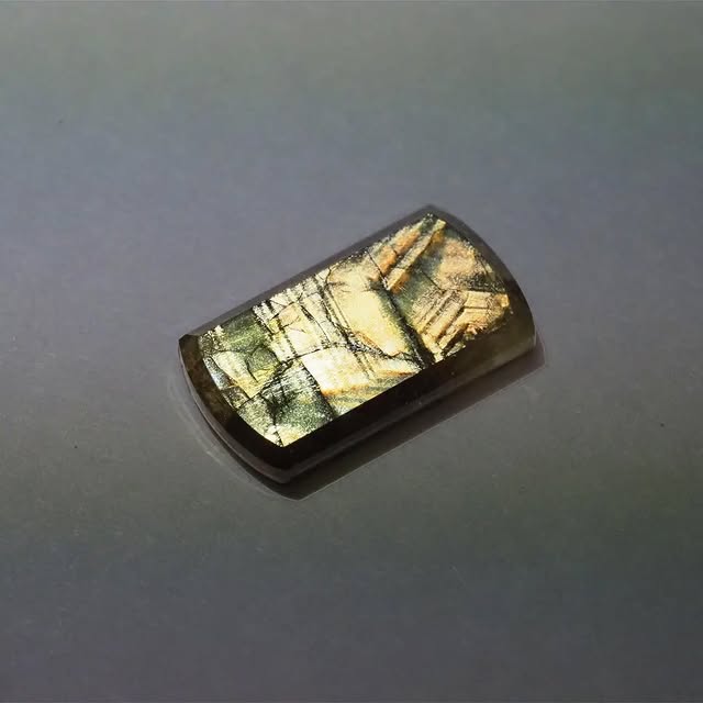 Polished gold sheen sapphire cabochon, rectangular cut, olive and gold tones, studio photograph on grey