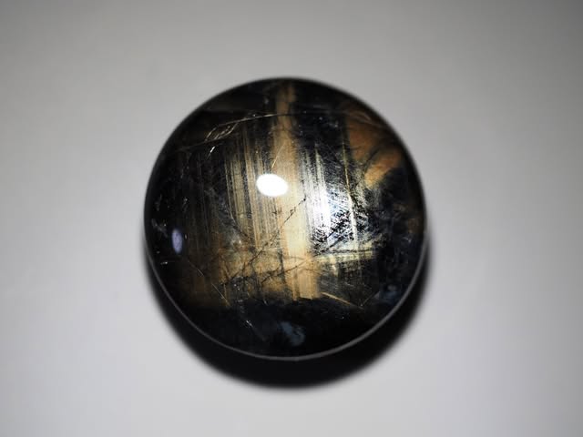 Round gold sheen sapphire cabochon with black and gold banding, studio photograph on grey